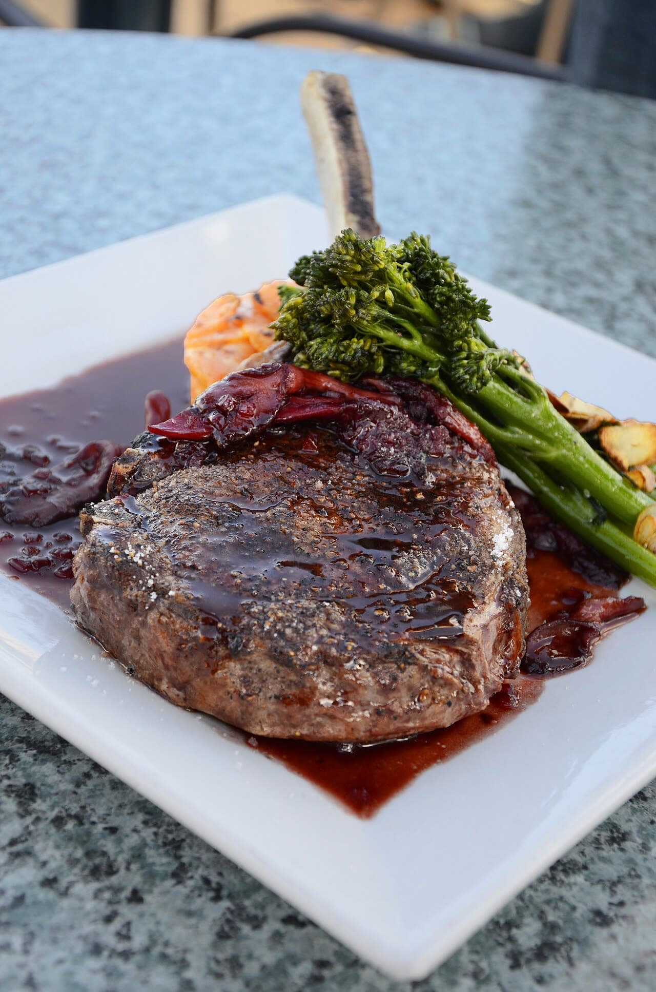 A thick bone-in steak on a white plate, glazed with dark sauce, a stalk of broccolini beside it, photographed close on a restaurant table.